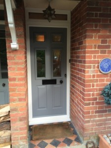 Grey front door framed by red brick, light and doormat