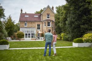 Rear view of mature couple looking at house. Man and woman are holding hands while standing on grassy field. They are wearing casuals.
